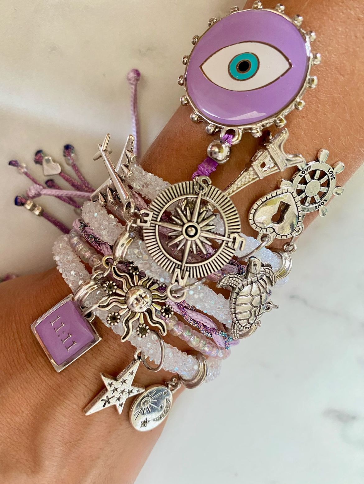 Weißes Strassarmband mit hellvioletten Perlen, violettem bösen Blick und Glücksbringern.
