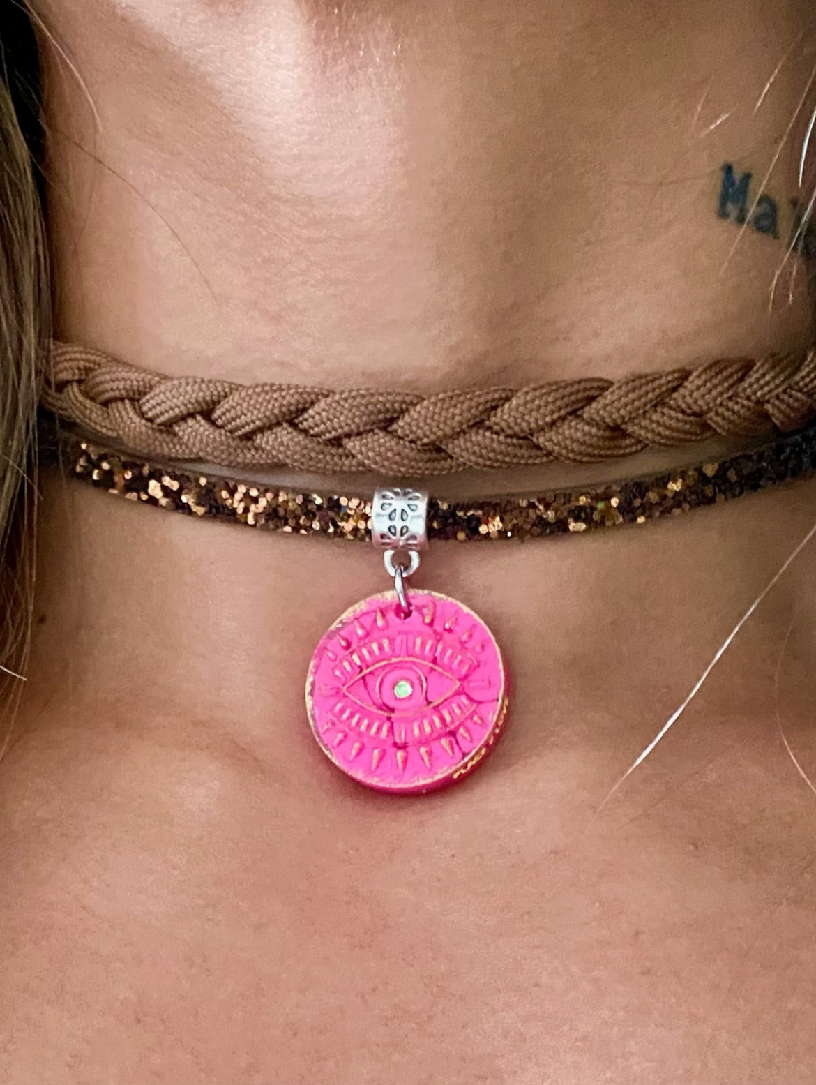 A close-up of a choker necklace worn on the neck, featuring a beige braided cord layered with a brown glitter strap. A circular pink pendant with a gold-engraved eye design and a small white center dot hangs at the center.