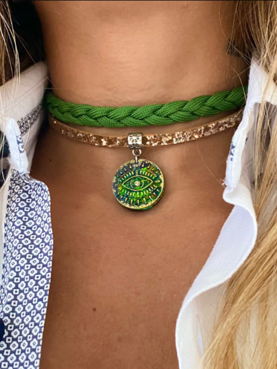 Layered choker necklace featuring a braided green cord and a gold glitter band, adorned with a green all-seeing eye pendant, styled on neck with an open collared shirt.