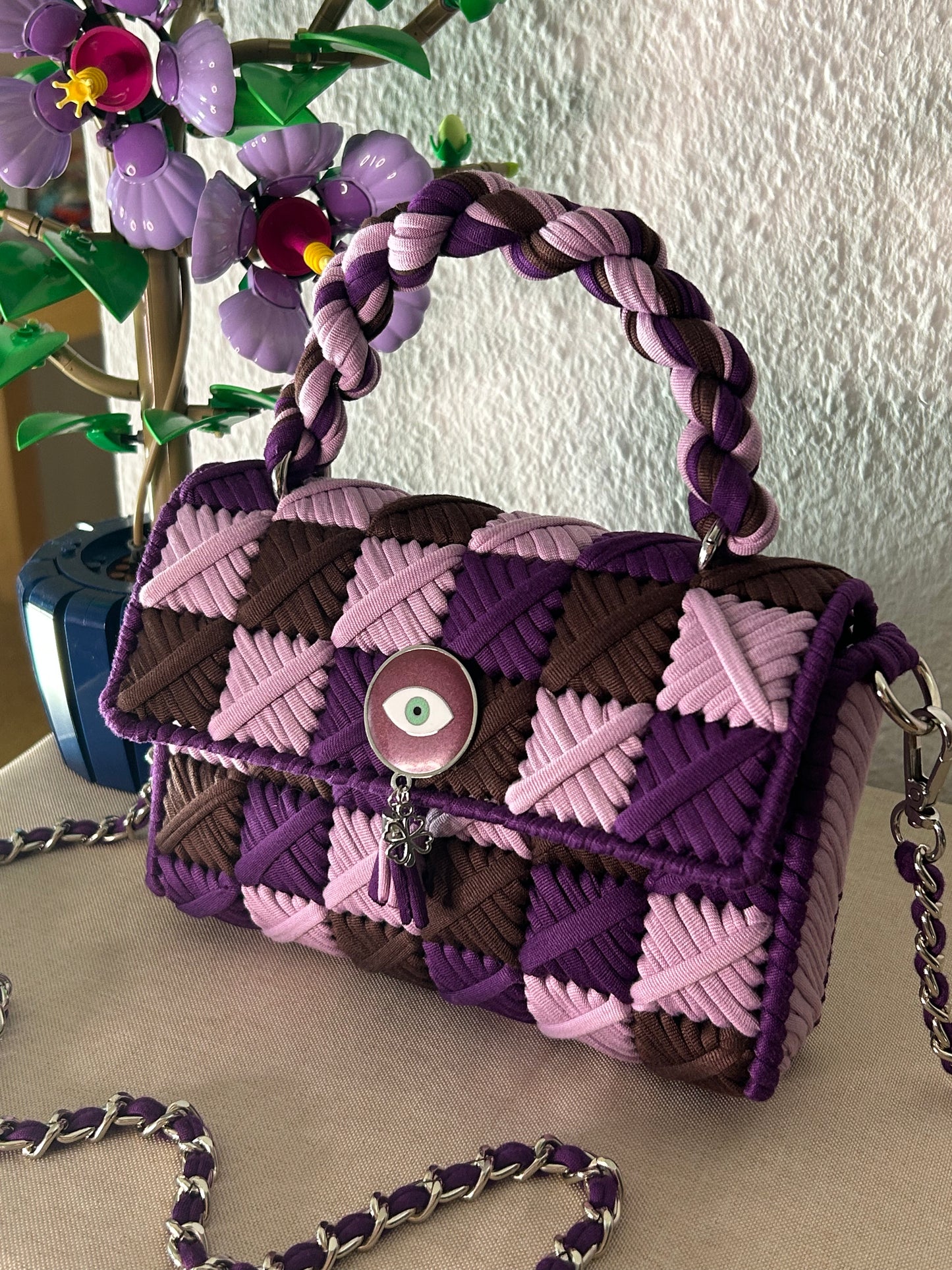 A small structured handbag with a braided top handle and chain strap, featuring a patchwork pattern in purple, pink, and dark brown. The front has a flap closure with a round eyelet charm and a tassel detail.