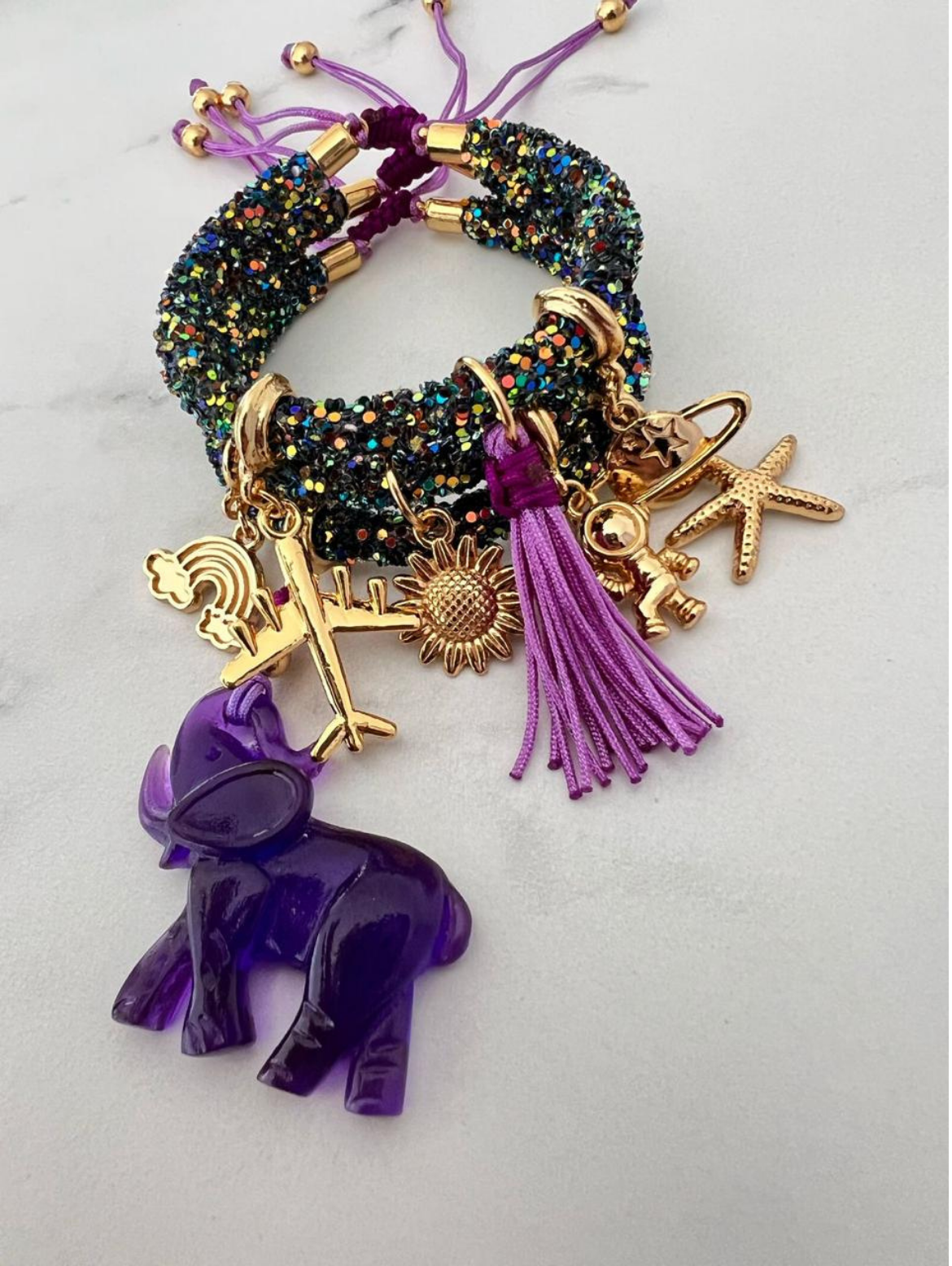 Stack of black glitter rhinestone cord bracelets with purple thread and gold-tone accents, featuring charms including a purple elephant, airplane, rainbow, sunflower, tassel, planet, astronaut, and starfish.