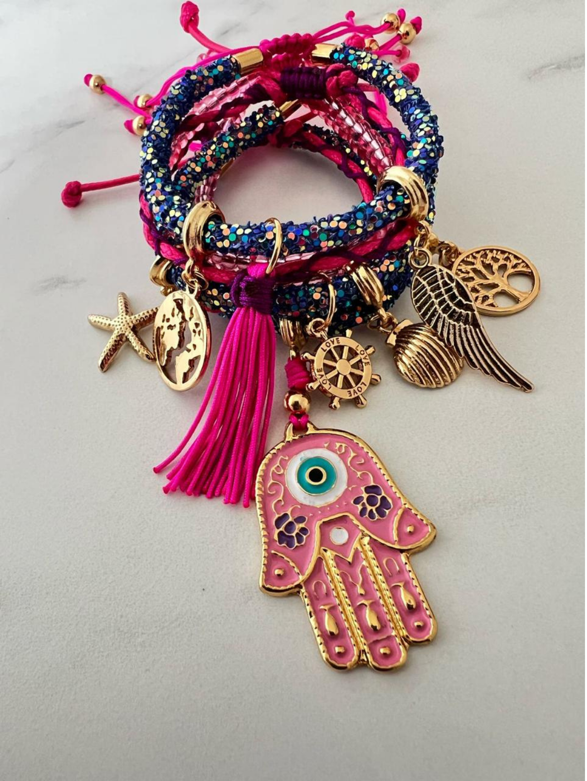 Stack of blue and pink rhinestone cord charm bracelets with fuchsia thread and gold-tone accents, featuring charms including a large pink Hamsa hand, tassel, world map, tree of life, angel wing, starfish, shell, compass, and 'LOVE' wheel