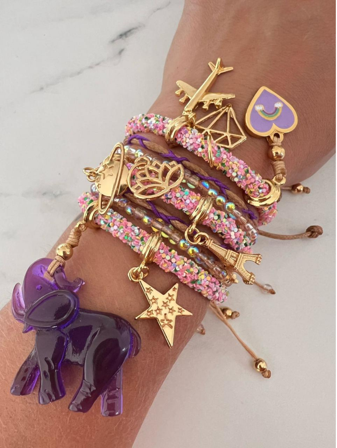 Stack of pastel pink rhinestone cord charm bracelets with gold accents, featuring charms including a purple elephant, airplane, planet, star, Eiffel Tower, lotus flower, geometric diamond, and a purple heart with rainbow.