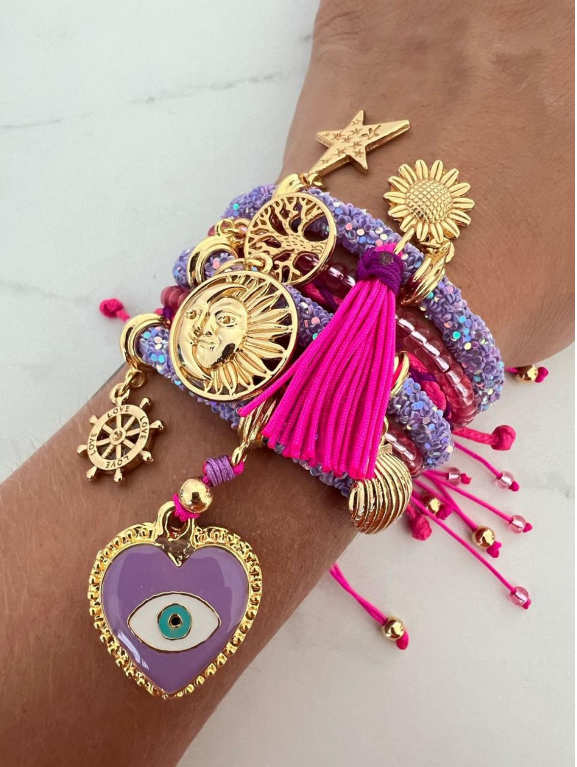 Stack of lavender and pink rhinestone cord charm bracelets with hot pink thread, featuring gold-tone charms including a purple evil eye heart, sun face, tree of life, sunflower, star, shell, tassel, and 'LOVE' compass wheel