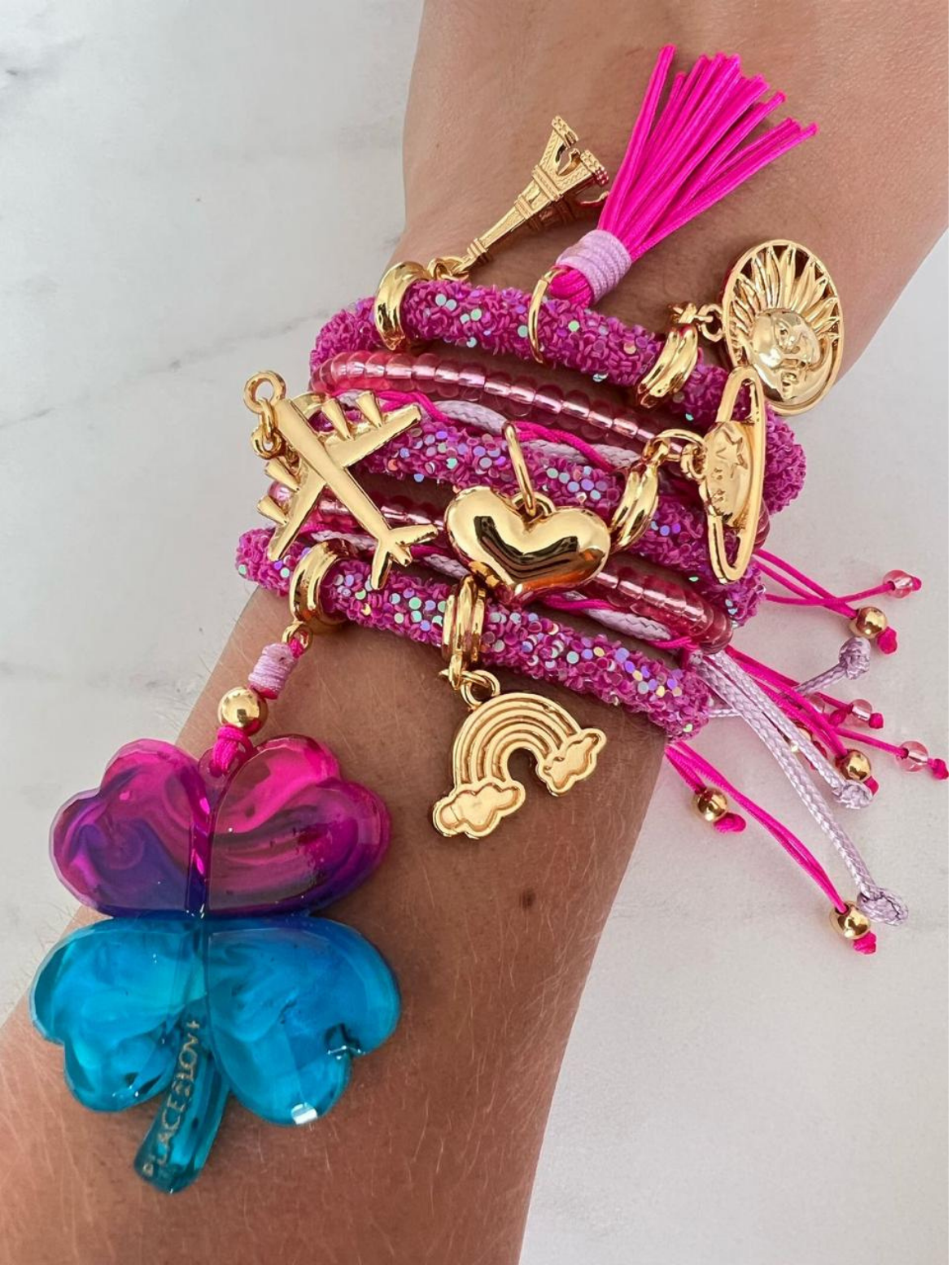 Stack of pink rhinestone cord charm bracelets with gold-tone accents and fuchsia thread, featuring charms including a large pink and blue four-leaf clover, airplane, heart, rainbow, Eiffel Tower, sun face, tassel, and planet.