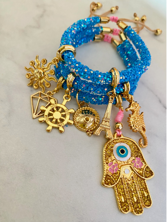 A sparkling blue glitter cord bracelet decorated with gold-tone charms, including a golden Hamsa hand with an evil eye and pink floral details, a sun, a diamond shape, a ship’s wheel, an all-seeing eye, the Eiffel Tower, and a seahorse. The bracelet is finished with gold hardware and beige adjustable cords.