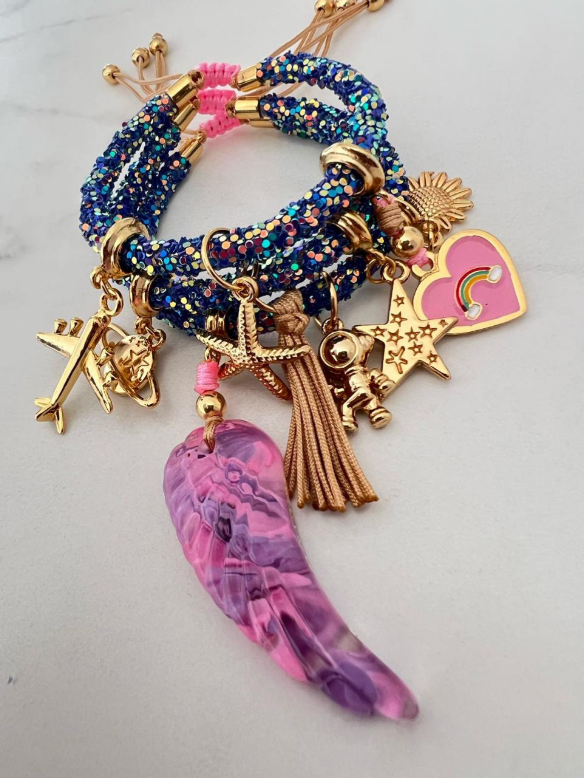 Stack of blue glitter rhinestone cord bracelets with pink thread and gold-tone accents, featuring charms including a pink and purple angel wing, airplane, starfish, astronaut, tassel, sunflower, and pink heart with rainbow.