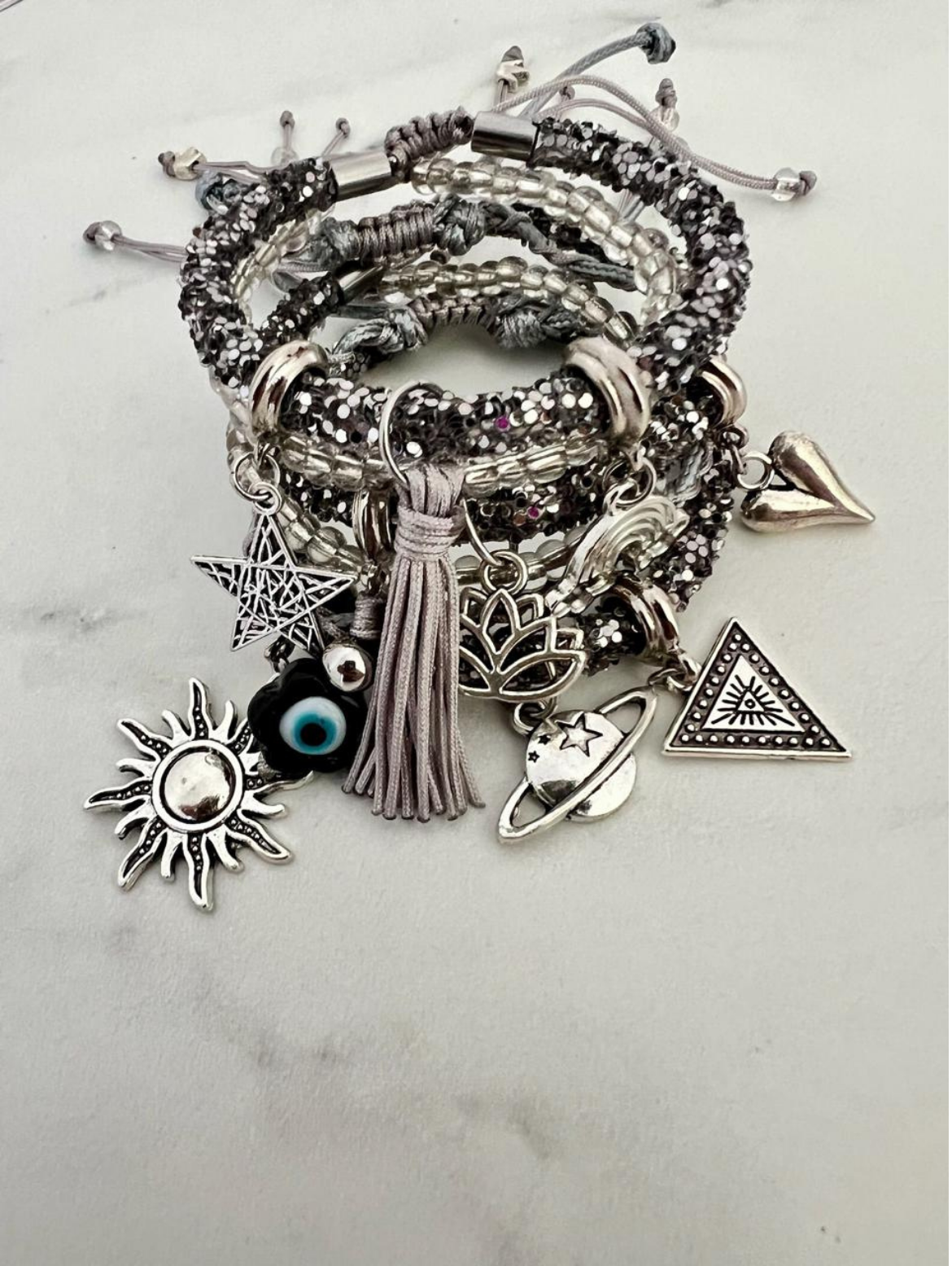 Stack of silver and grey rhinestone cord charm bracelets with clear beads and grey tassels, featuring silver charms including a sun, star, evil eye bead, lotus flower, triangle symbol, heart, and planet with ring.