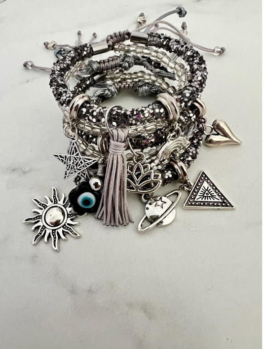 Stack of silver and grey rhinestone cord charm bracelets with clear beads and grey tassels, featuring silver charms including a sun, star, evil eye bead, lotus flower, triangle symbol, heart, and planet with ring.