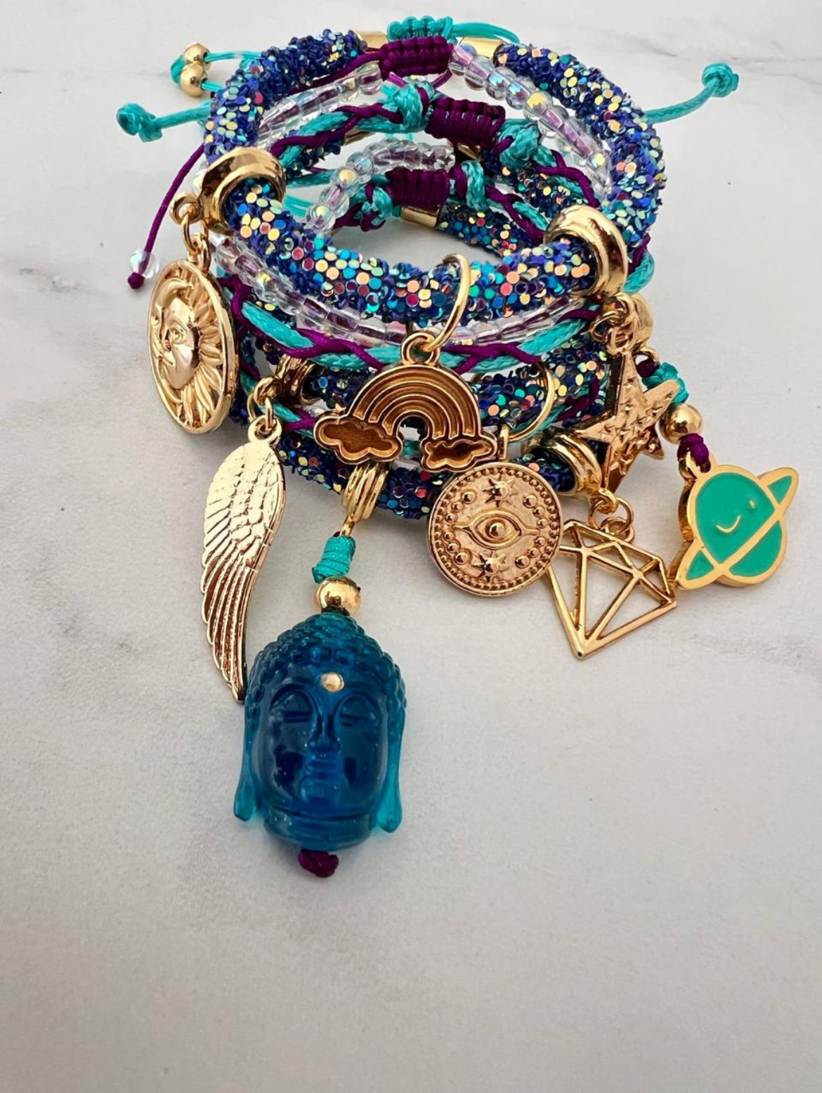 Stack of blue and purple rhinestone cord bracelets with turquoise and gold accents, featuring charms including a blue Buddha head, sun face, rainbow, angel wing, all-seeing eye, planet, star, and geometric diamond.