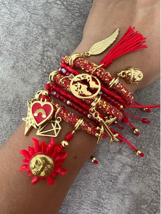 Stack of red rhinestone cord charm bracelets with gold accents, featuring gold-tone and enamel charms including a world map, airplane, heart with rainbow, diamond outline, shooting star, angel wing, tassel, and a red sun with gold face