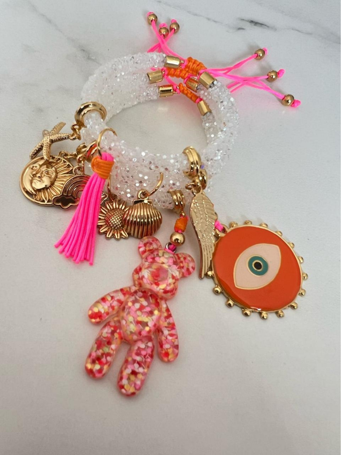 Stack of white rhinestone cord charm bracelets with neon pink and orange thread, featuring gold-tone charms including a large orange evil eye medallion, pink confetti bear, angel wing, tassel, sun face, rainbow, sunflower, shell, and starfish.