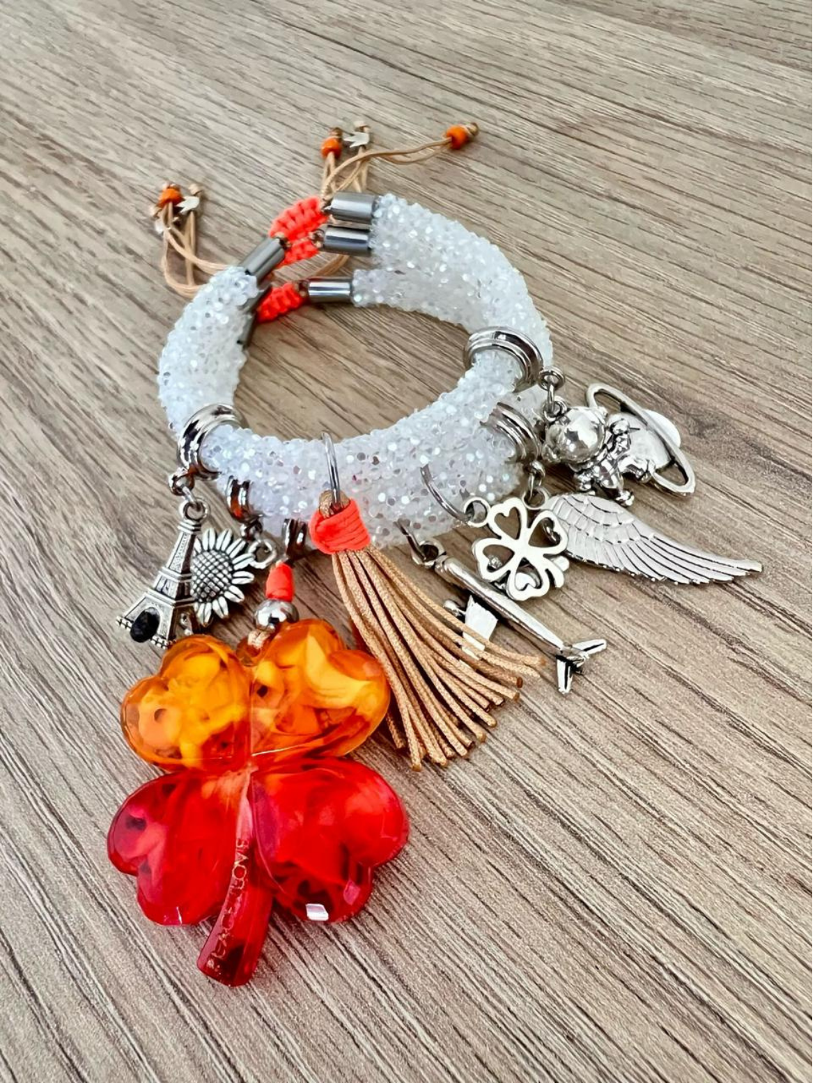 Stack of rhinestone cord charm bracelets with orange thread accents, featuring silver charms and a large red-orange acrylic four-leaf clover, tassel, angel wing, Eiffel Tower, and astronaut