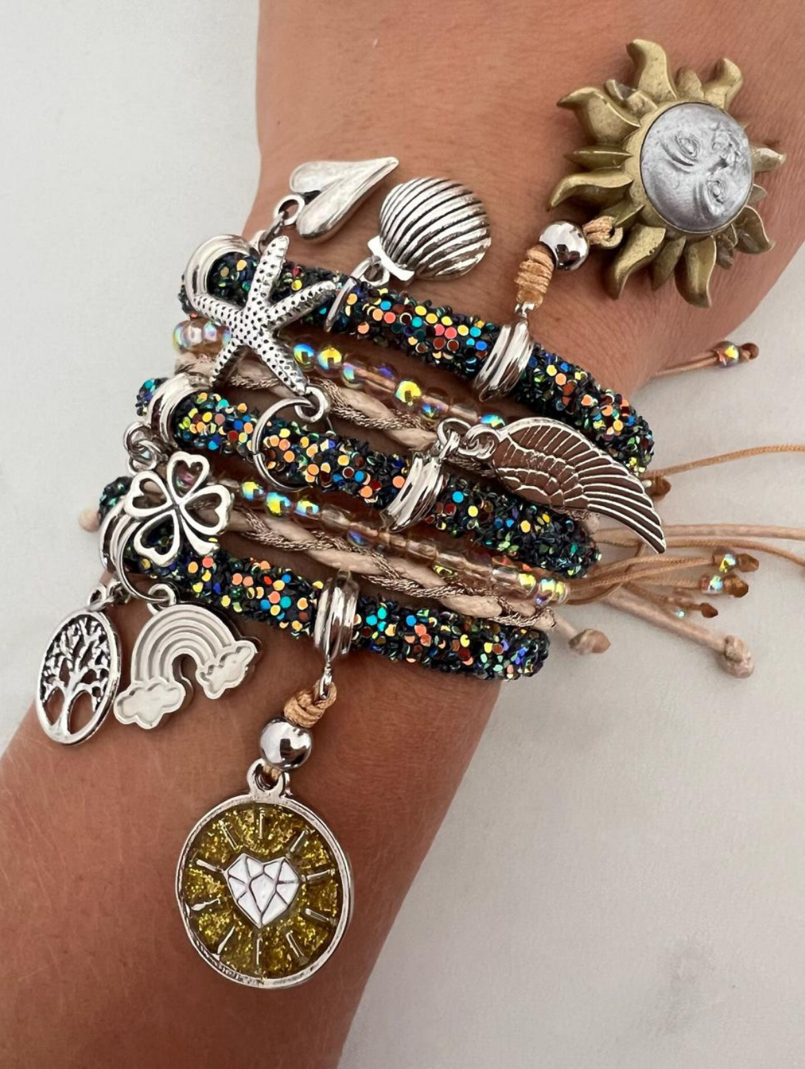 Stack of glittery black and beige charm bracelets on a wrist, decorated with silver and gold charms including a sun face, heart, angel wing, clover, rainbow, tree of life, shell, and starfish