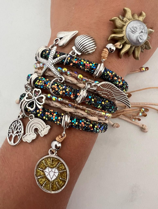 Stack of glittery black and beige charm bracelets on a wrist, decorated with silver and gold charms including a sun face, heart, angel wing, clover, rainbow, tree of life, shell, and starfish