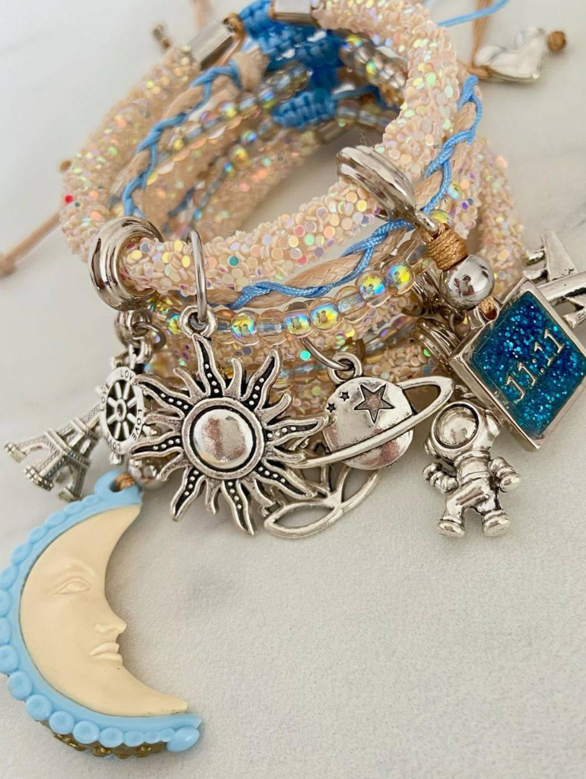 Stack of handmade beaded and woven charm bracelets with celestial and travel-themed silver charms including moon, sun, astronaut, aeroplane, planet, and Eiffel Tower on a white background.”