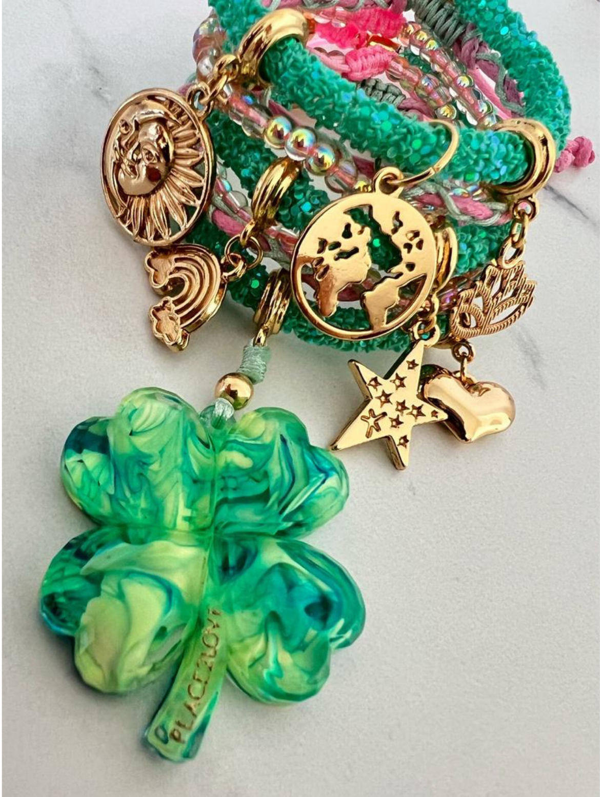 Stack of green and pink rhinestone cord charm bracelets with gold-tone accents, featuring charms including a large marbled green four-leaf clover, sun face, world map, rainbow, angel wing, shooting star, and gold heart.