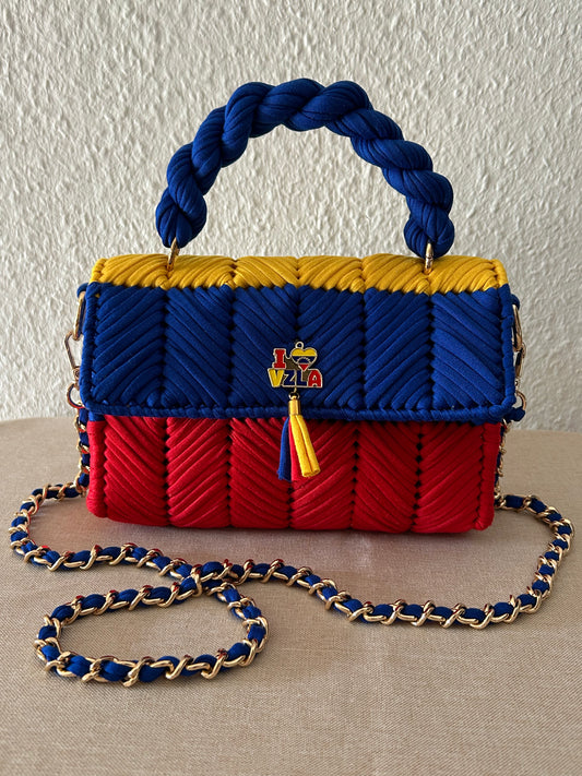 A woven handbag with a braided top handle and chain strap, featuring horizontal panels in blue, yellow, and red, with a small multicolor logo charm on the front.