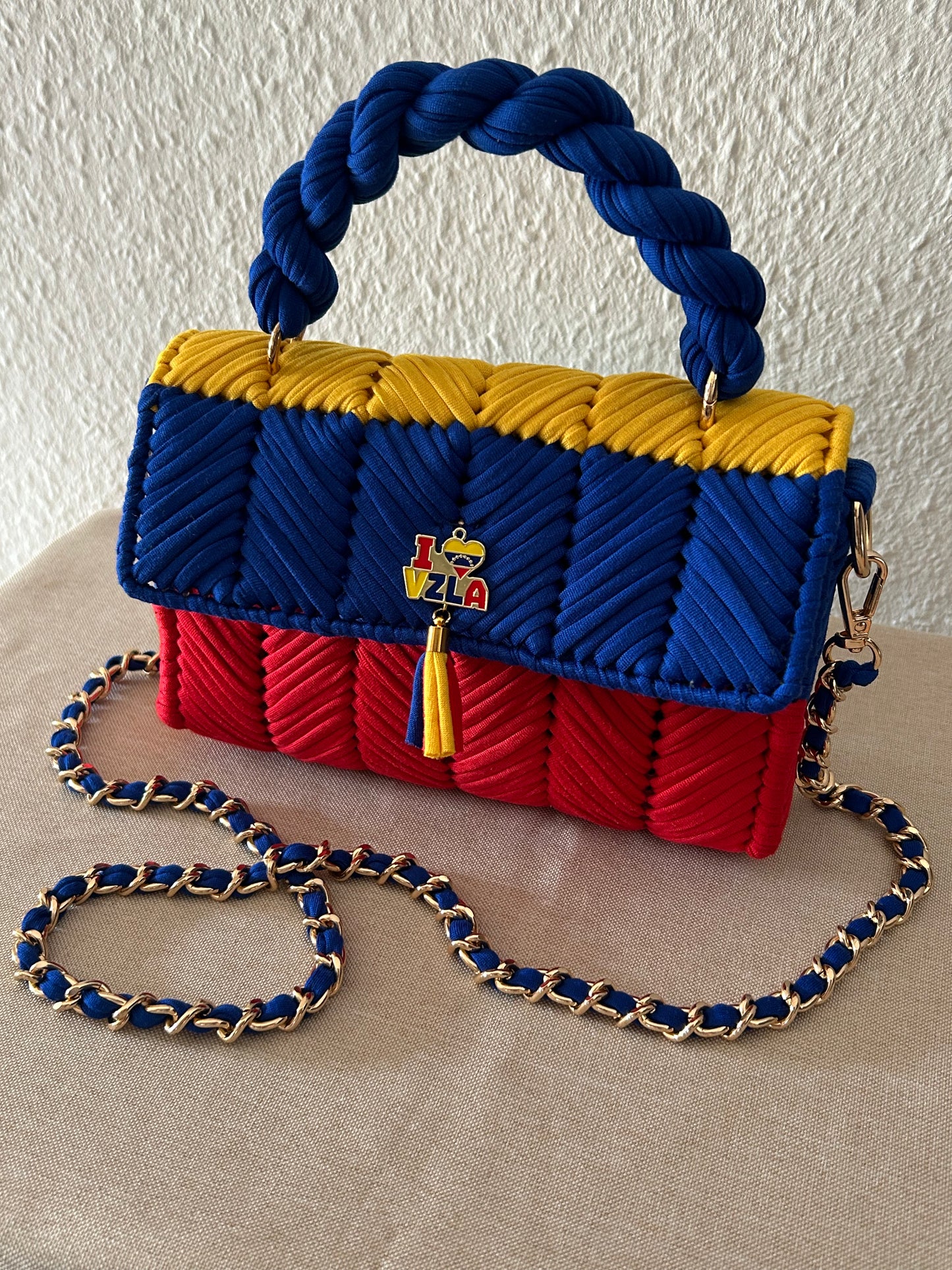 A woven handbag with a braided top handle and chain strap, featuring horizontal bands of blue, yellow, and red. A small colorful logo charm is centered on the front flap, and the bag is displayed on a beige fabric surface against a textured off-white wall.