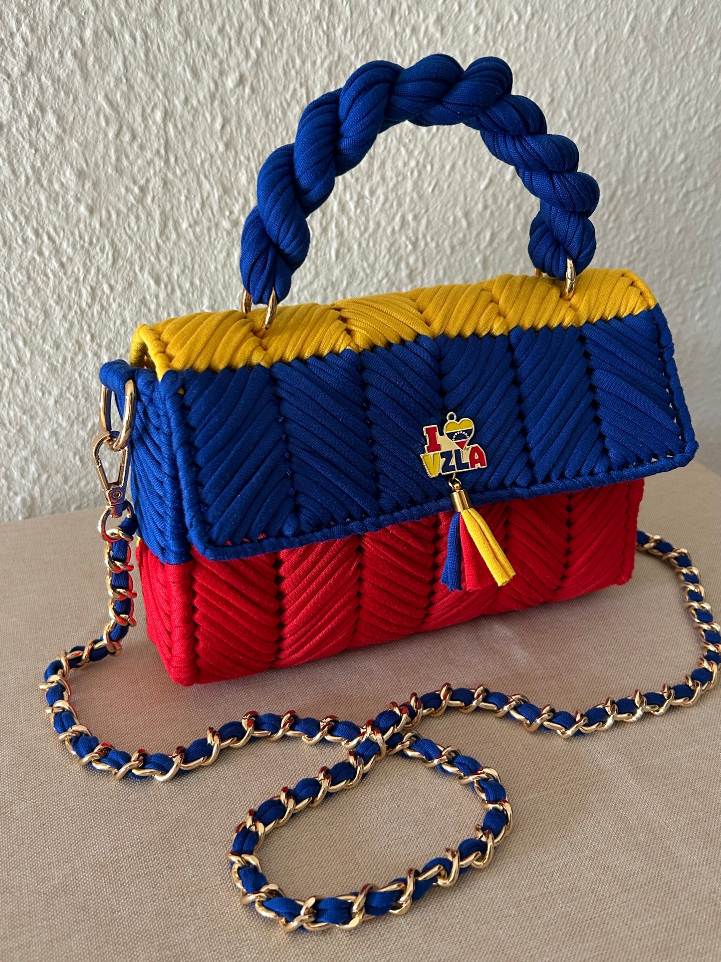 A woven handbag with a braided rope handle and a gold-tone chain strap, featuring horizontal bands of blue, yellow, and red. A small 'I <3 ZLA' pin is attached to the front flap, set against a neutral textured wall and beige surface.