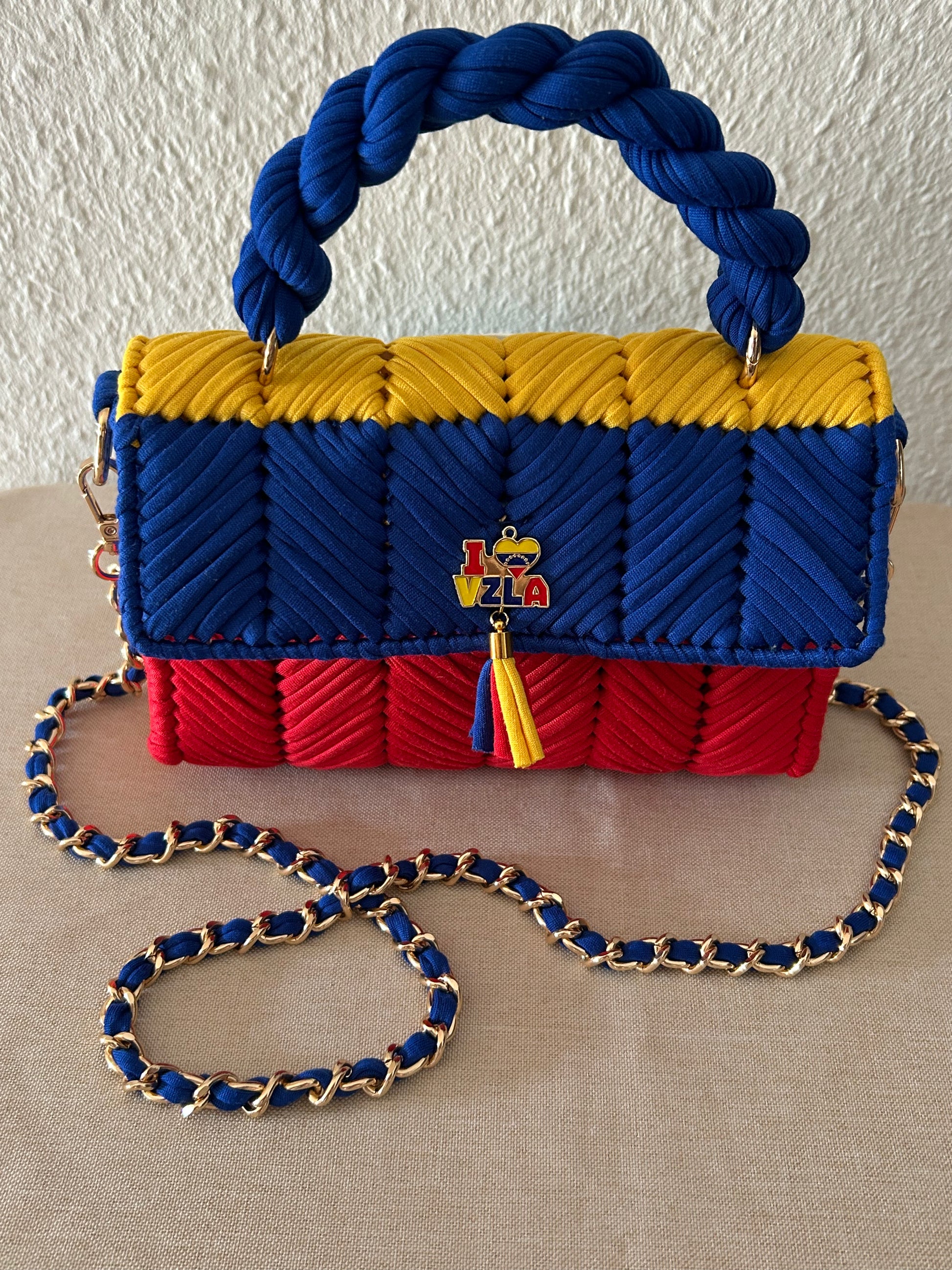 A woven handbag with a braided rope handle and chain strap, featuring horizontal stripes of blue, yellow, and red, with a small colorful 'VIVA' tassel charm in the center.