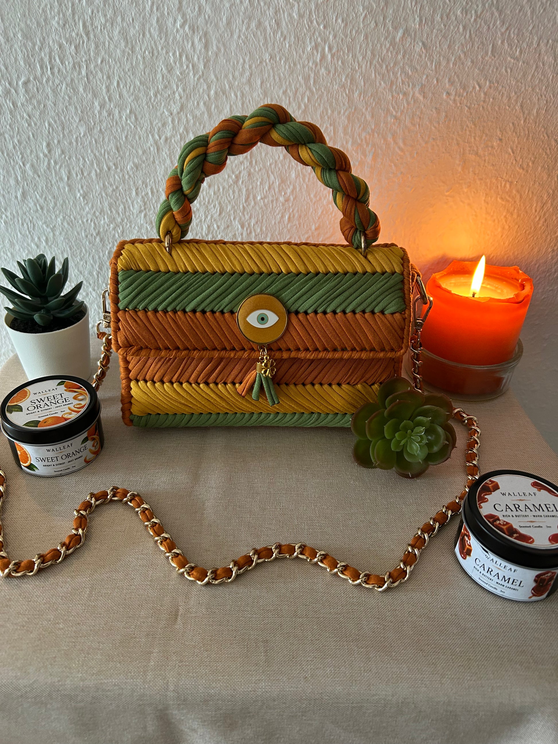 A woven rectangular handbag with a braided top handle sits on a beige fabric-covered surface. The bag has horizontal woven stripes in yellow, green, and orange-brown, with a round gold-tone front clasp and a dangling tassel charm.