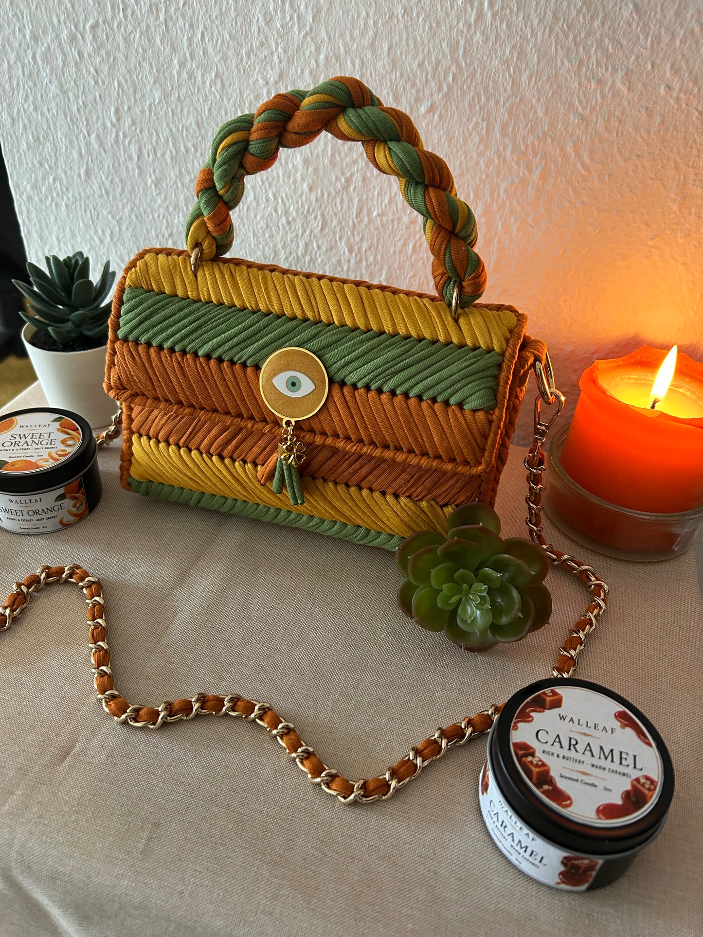 A woven handbag with a braided top handle in yellow, green, and orange-brown, styled on a beige fabric surface with a gold chain strap, succulents, and a lit candle.