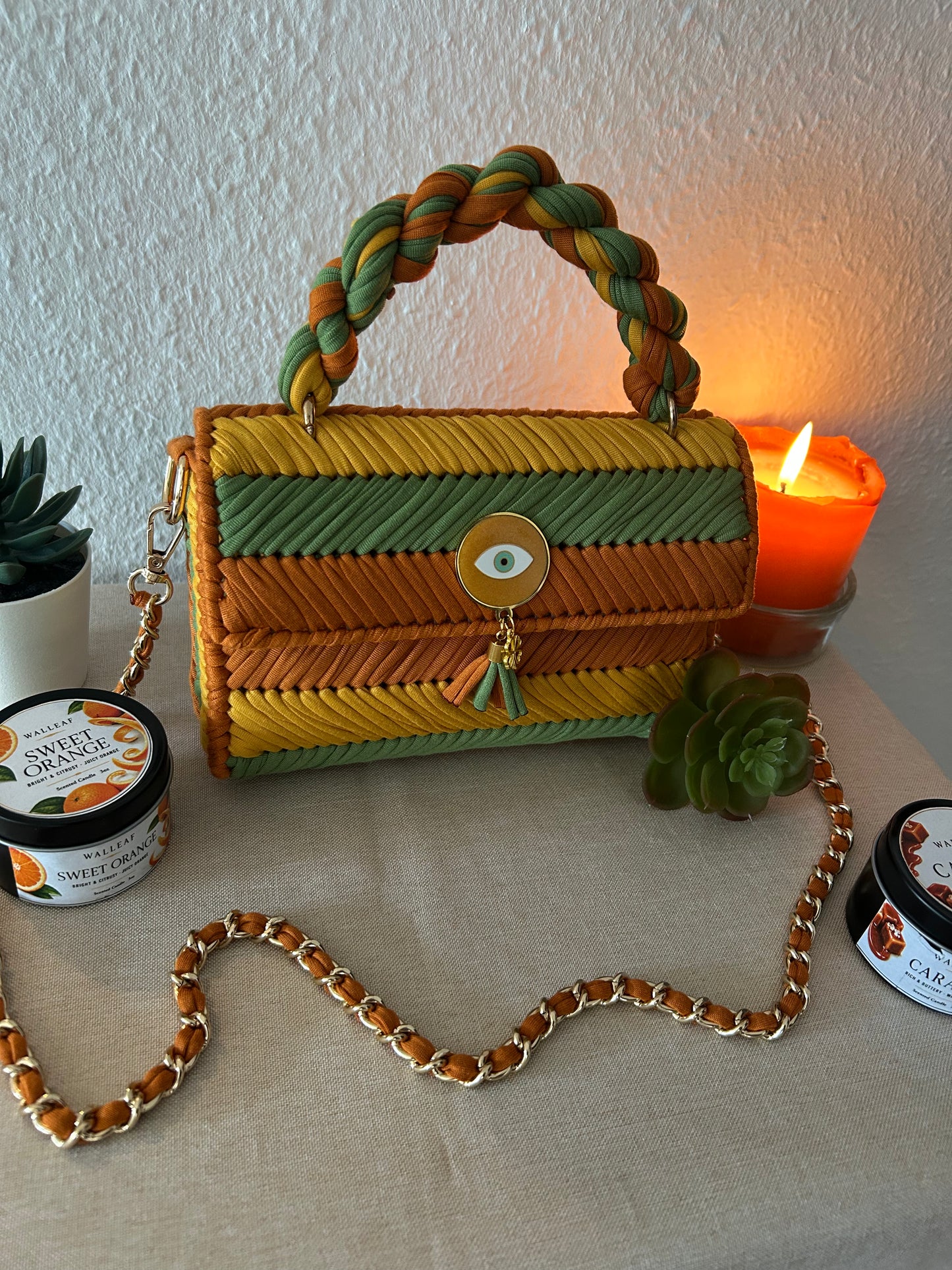A woven handbag with a braided top handle, featuring horizontal stripes in yellow, green, and orange-brown. It has a gold-tone chain strap, a tassel charm with an eye motif, and is styled on a beige fabric surface with a lit orange candle, succulents, and decorative jars.