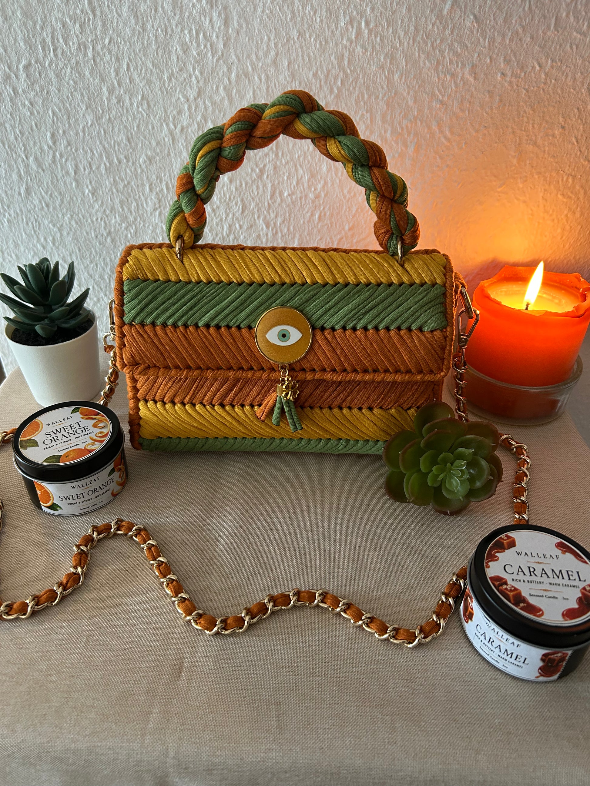 A woven handbag with a braided top handle, featuring horizontal stripes in yellow, orange, and green, and a gold-tone eye-shaped clasp. The bag is styled on a beige fabric surface with a gold chain strap, flanked by candles, succulents, and two jars of 'The Art of Living' Caramel soap.