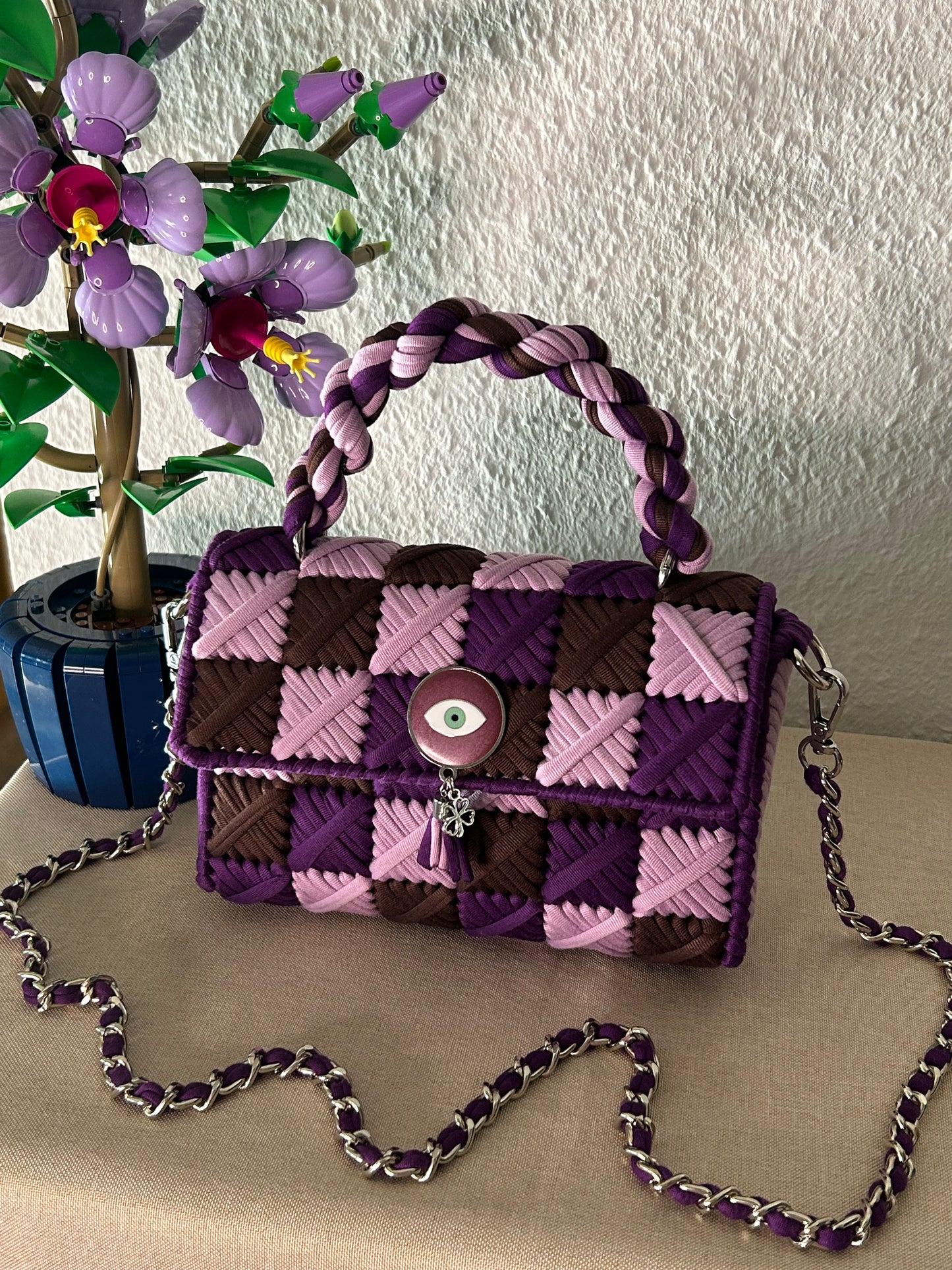 A purple and pink woven handbag with a braided top handle and silver chain strap, displayed on a beige surface next to a decorative plant.