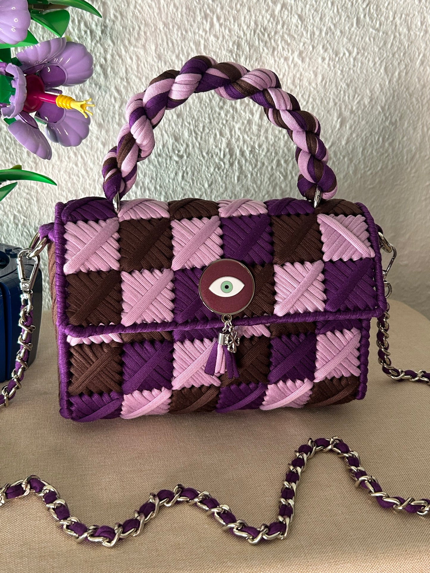 A purple and pink woven leather handbag with a braided top handle, a silver chain strap, and a central evil eye clasp, displayed on a beige surface against a textured wall.