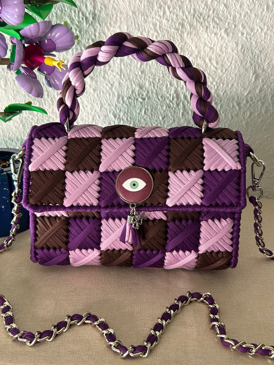 A woven leather handbag with a purple, pink, and brown checkered pattern, featuring a braided top handle, a silver chain strap, and a central evil eye clasp.