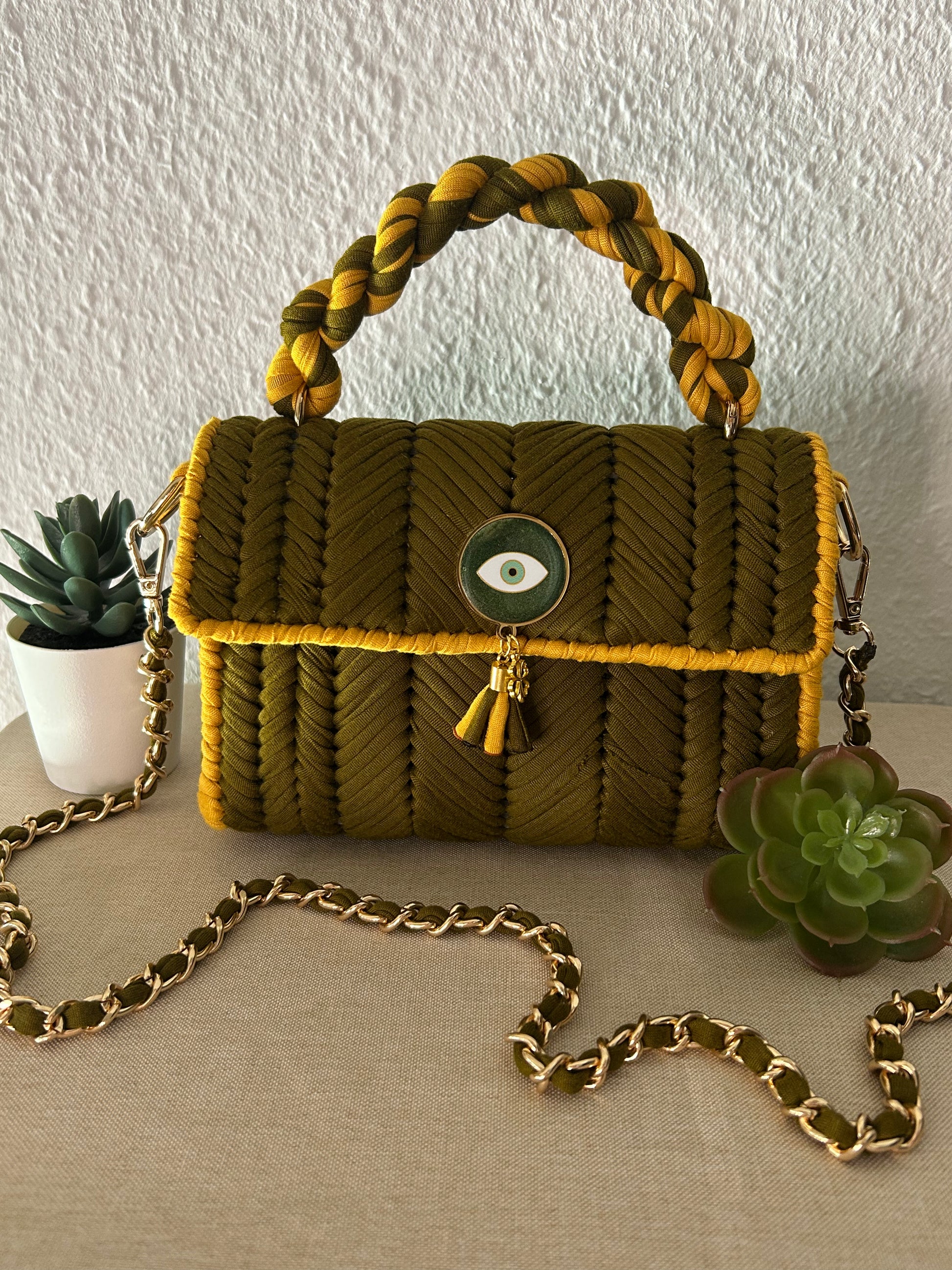 An olive green and mustard yellow braided handbag with a braided top handle, a green evil eye charm, and a gold chain strap, styled on a beige fabric surface with succulent plants.