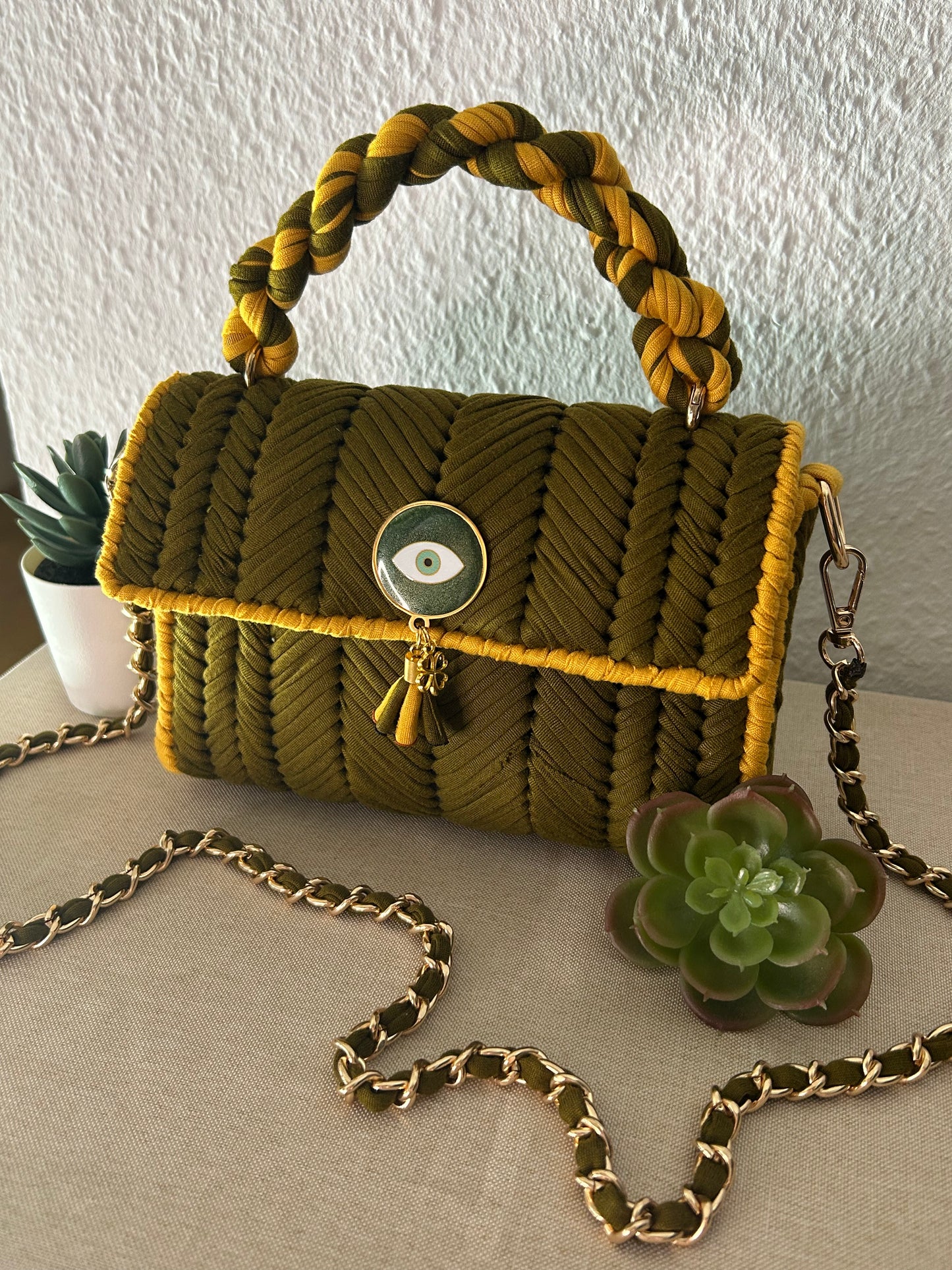 A woven olive green and mustard yellow handbag with a braided top handle, a gold-tone chain strap, and a decorative evil eye clasp, styled on a neutral surface with succulent plants.