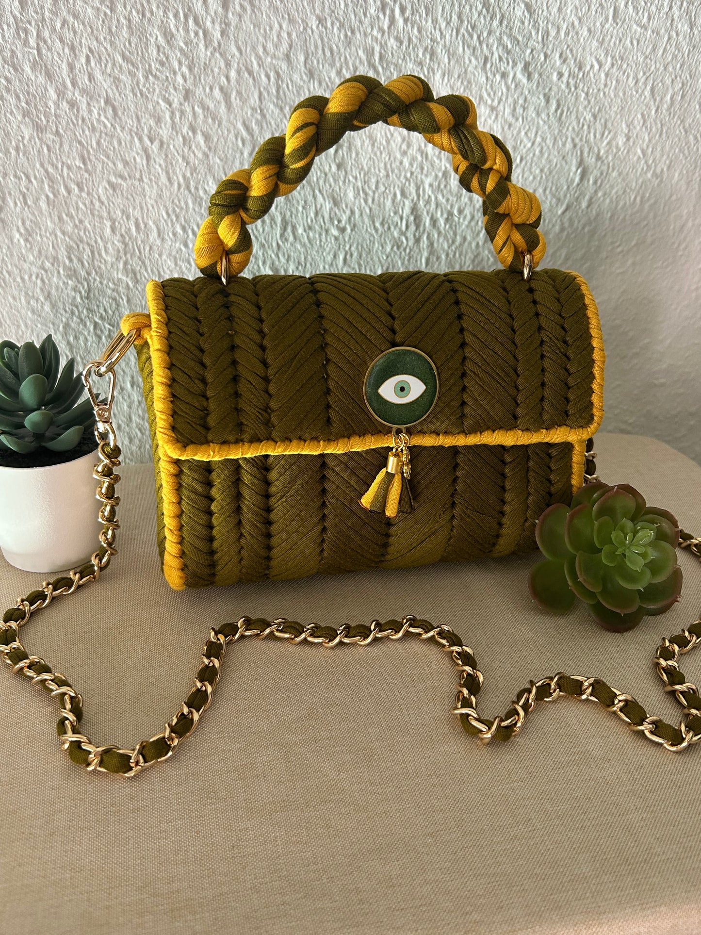A textured olive green and mustard yellow braided handbag with a braided top handle, gold-tone chain strap, and a green evil eye button closure, styled on a beige fabric surface with succulent plants.